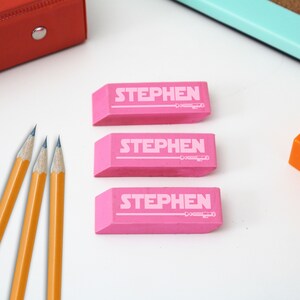 以下が含まれることがあります： 「STEPHEN」という名前が白で印刷された、ピンク色の長方形の消しゴムが3つあります。