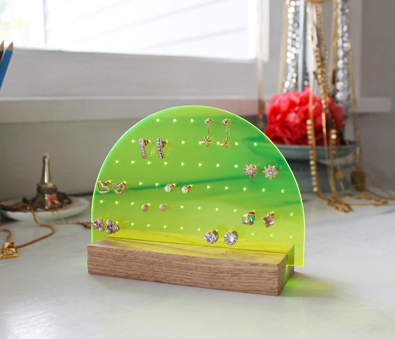 May include: A green and yellow semi-circle earring holder with a wooden base. The holder has many small holes to hold earrings. There are many different earrings on the holder, including gold, silver, and pearl earrings.