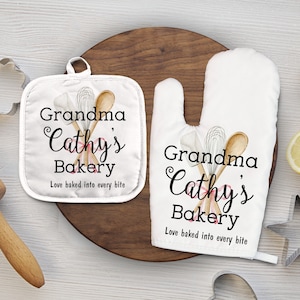 Puede incluir: Un juego de dos guantes de horno blancos con una cinta rosa y utensilios de cocina de madera. Los guantes tienen el texto "Grandma Cathy's Bakery Love baked into every bite" impreso en ellos.