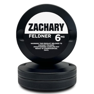 以下が含まれることがあります： 白いラベルに「ZACHARY FELDNER」と「6MG」と印刷された黒い円形製品。製品はテクスチャ加工されたベースを持ち、不正な取り扱いに関する警告が含まれています。