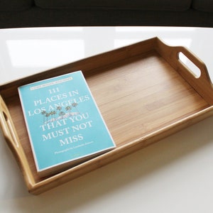 May include: A wooden serving tray with a book titled "111 Places in Los Angeles That You Must Not Miss" on top. The tray has two handles on the sides.