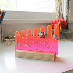 May include: A pink acrylic jewelry organizer with a wooden base. The organizer has multiple slots for rings and a grid of holes for earrings. The organizer is filled with various rings and earrings.