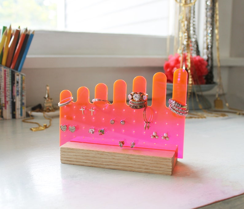 May include: A pink acrylic jewelry organizer with a wooden base. The organizer has multiple slots for rings and a grid of holes for earrings. The organizer is filled with various rings and earrings.