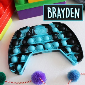 Puede incluir: Un juguete pop it fidget azul y negro con forma de mando de videojuego. El juguete tiene el nombre "Brayden" escrito en él.