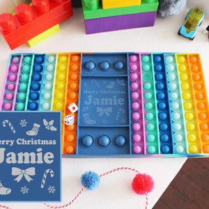 May include: A rainbow pop-it fidget toy with a blue frame and a personalized message that says "Merry Christmas Jamie".
