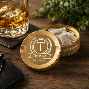 May include: A round, gold-colored metal tin with a textured edge, featuring a white engraved design on the lid. The design includes a central letter T inside a circle, flanked by laurel branches, with the name THOMAS and EST. 11.10.2026 below.