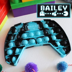 May include: A blue and black pop it fidget toy shaped like a video game controller. The toy has a small screen that says "BAILEY" with a Pac-Man character on either side.