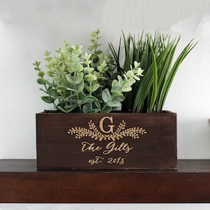 Puede incluir: Una jardinera de madera marrón oscuro con el texto "The Gill's est. 2015" y la letra "G" grabada en la parte delantera. La jardinera está llena de plantas verdes.
