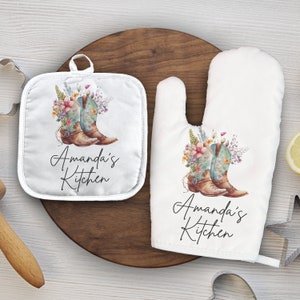 May include: A set of two white cotton oven mitts with a floral design and cowboy boots. The text "Amanda's Kitchen" is printed on both mitts.