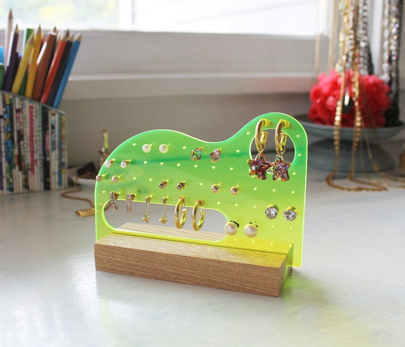 May include: A green and yellow acrylic earring holder with a wooden base. The holder has multiple holes for earrings and is filled with various gold and pearl earrings.