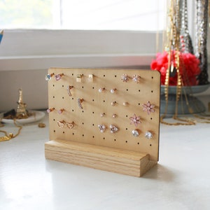 May include: A wooden earring display stand with a light brown finish. The stand has a pegboard with multiple holes for holding earrings. The pegboard is filled with a variety of earrings, including gold, silver, and pink.
