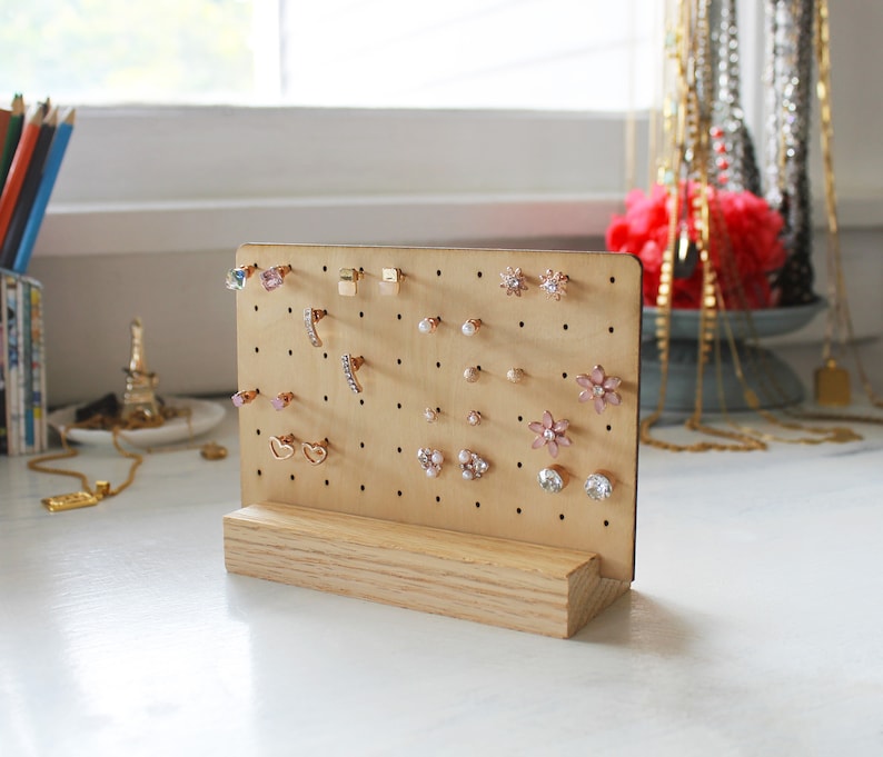 May include: A wooden earring display stand with a light brown finish. The stand has a pegboard with multiple holes for holding earrings. The pegboard is filled with a variety of earrings, including gold, silver, and pink.