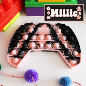 Puede incluir: Un juguete pop it de color rosa y negro con forma de mando de videojuego. El juguete tiene el nombre "Millie" escrito en él.