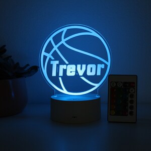 Pu&ograve; includere: Una luce notturna personalizzata a tema basket con il nome "Trevor" illuminato in bianco. La luce &egrave; blu e poggia su una base bianca, con un telecomando nelle vicinanze. Sullo sfondo c'&egrave; una pianta decorativa.