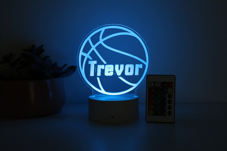 Pu&ograve; includere: Una luce notturna personalizzata a tema basket con il nome "Trevor" illuminato in bianco. La luce &egrave; blu e poggia su una base bianca, con un telecomando nelle vicinanze. Sullo sfondo c'&egrave; una pianta decorativa.