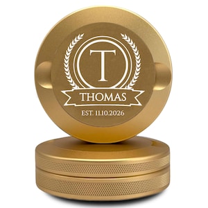 May include: A gold-colored ashtray with a circular design. The top features a white emblem with the letter "T" inside a wreath, the name "Thomas", and the date "EST. 11.10.2026". The ashtray has a textured base and a small notch for resting items.