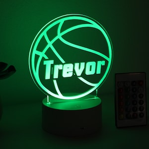 Pu&ograve; includere: Luce notturna verde a tema basket con il nome "Trevor" illuminato in bianco. La luce &egrave; su una base bianca, con un telecomando a destra. Il tema &egrave; sport e personalizzazione.