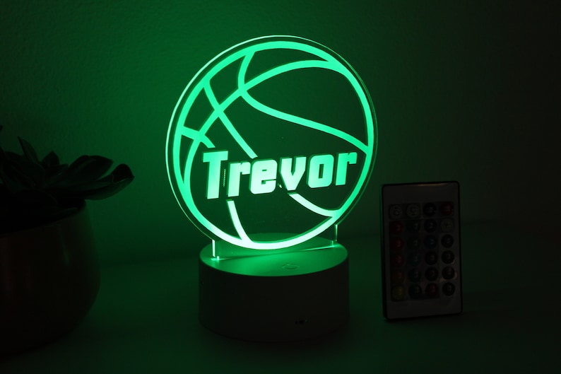 Pu&ograve; includere: Luce notturna verde a tema basket con il nome "Trevor" illuminato in bianco. La luce &egrave; su una base bianca, con un telecomando a destra. Il tema &egrave; sport e personalizzazione.