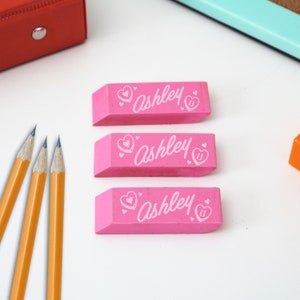 May include: Three pink rectangular erasers with the name "Ashley" and a heart design printed on each eraser.