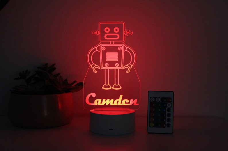 May include: A red LED night light shaped like a robot with the name "Camden" underneath. The light is on a white base and comes with a remote control.