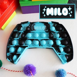 May include: A blue and black pop it fidget toy shaped like a video game controller. The toy has a label that says "MILO" on it.