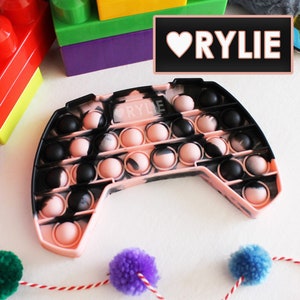 May include: A pink and black pop it fidget toy shaped like a video game controller. The toy has a heart with the name "RYLIE" written inside.