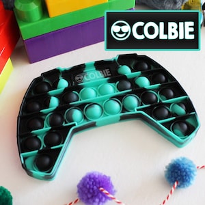 Puede incluir: Un juguete pop-it con forma de mando negro y verde azulado con la palabra "COLBIE" impresa. El juguete tiene múltiples círculos con forma de burbuja que se pueden empujar hacia adentro y hacia afuera. El fondo incluye bloques de construcción coloridos y pompones.