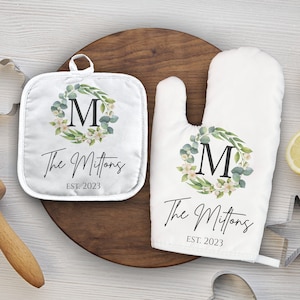 Puede incluir: Un juego de agarraderas y guantes para horno blancos con un diseño de corona floral y la letra "M" en el centro. El texto "The Mittons Est. 2023" está impreso debajo de la corona.
