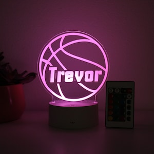 Pu&ograve; includere: Una luce notturna a tema basket con il nome "Trevor" illuminato in bianco. La luce &egrave; posta su una base bianca, con un telecomando visibile. La luce emette un morbido bagliore rosa.
