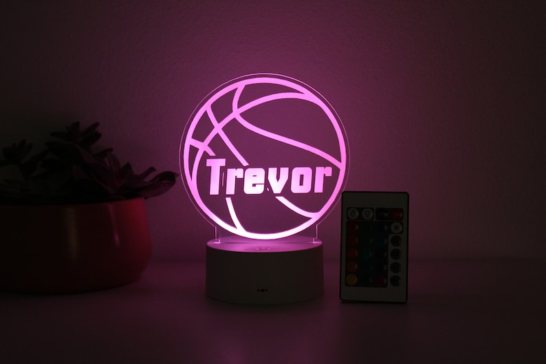 Pu&ograve; includere: Una luce notturna a tema basket con il nome "Trevor" illuminato in bianco. La luce &egrave; posta su una base bianca, con un telecomando visibile. La luce emette un morbido bagliore rosa.