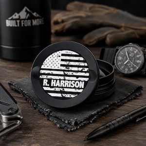May include: A black circular metal grinder featuring a white distressed American flag graphic and the text R. HARRISON printed on the lid. The object is positioned on a dark fabric square, surrounded by a black pen, a watch, and a metal container.