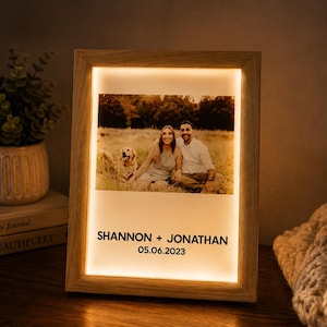 May include: A rectangular light-up picture frame with a light wood finish. The frame displays a photograph of two people and a dog sitting in a field, with the text SHANNON + JONATHAN and the date 05.06.2023 printed in black below the image.