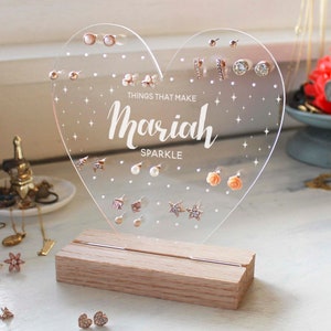 May include: Clear acrylic heart-shaped earring holder with a wooden base. The holder has a white inscription that reads "Things that make Mariah Sparkle".