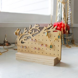 May include: A wooden jewelry stand shaped like a tree branch with a light wood finish. The stand has multiple holes for earrings and hooks for necklaces. There are several pairs of earrings and necklaces on the stand.