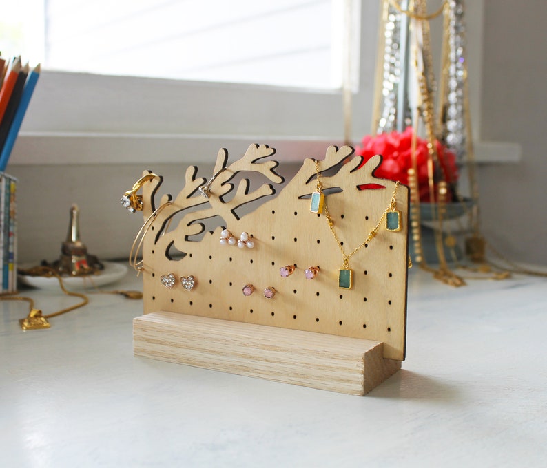 May include: A wooden jewelry stand shaped like a tree branch with a light wood finish. The stand has multiple holes for earrings and hooks for necklaces. There are several pairs of earrings and necklaces on the stand.
