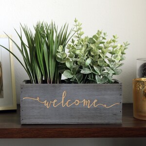 以下が含まれることがあります： グレーの木製のプランターボックスに、金色の文字で「welcome」と刻まれています。プランターには緑の葉の植物が植えられています。
