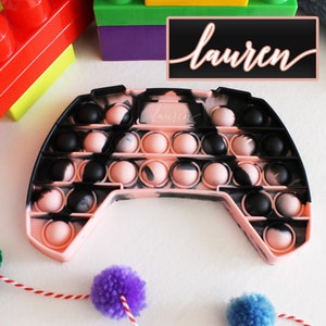 Puede incluir: Juguete pop it de silicona rosa y negro con forma de mando de videojuego. El juguete tiene el nombre "lauren" escrito en él.