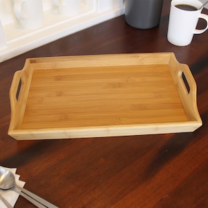 May include: A rectangular bamboo serving tray with two handles on the sides. The tray is empty and sitting on a wooden surface.