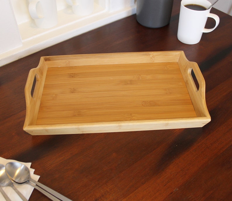 May include: A rectangular bamboo serving tray with two handles on the sides. The tray is empty and sitting on a wooden surface.