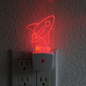 May include: A white night light shaped like a rocket ship with the name "Adam" printed on it. The night light is plugged into a white electrical outlet and is glowing red.