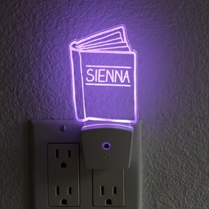 May include: A purple night light shaped like an open book with the word "SIENNA" written on the cover. The night light is plugged into a white electrical outlet.