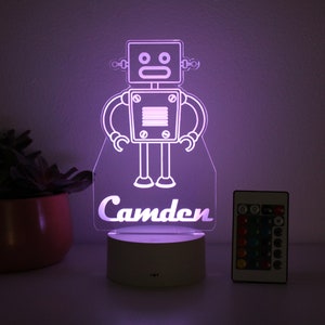 May include: A white robot night light with a purple glow, featuring the name "Camden" at the bottom. The light comes with a remote control.