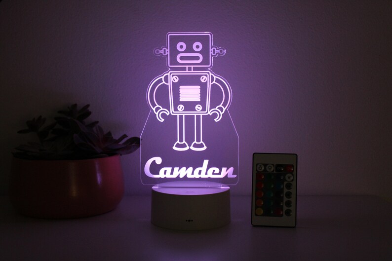 May include: A white robot night light with a purple glow, featuring the name "Camden" at the bottom. The light comes with a remote control.
