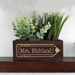 May include: A dark brown wooden planter shaped like a pencil with the text "Mrs. Richland" engraved on the front. The planter is filled with green plants.