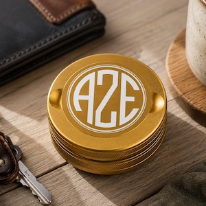 May include: A circular gold-colored metal grinder featuring a white monogram with the letters AZE centered on the top lid. The exterior of the lid has a textured, knurled edge for grip, and the surface is smooth with two small indentations.