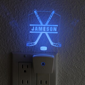 May include: A white hockey stick night light with the name "JAMESON" printed on it. The light is plugged into a white electrical outlet.