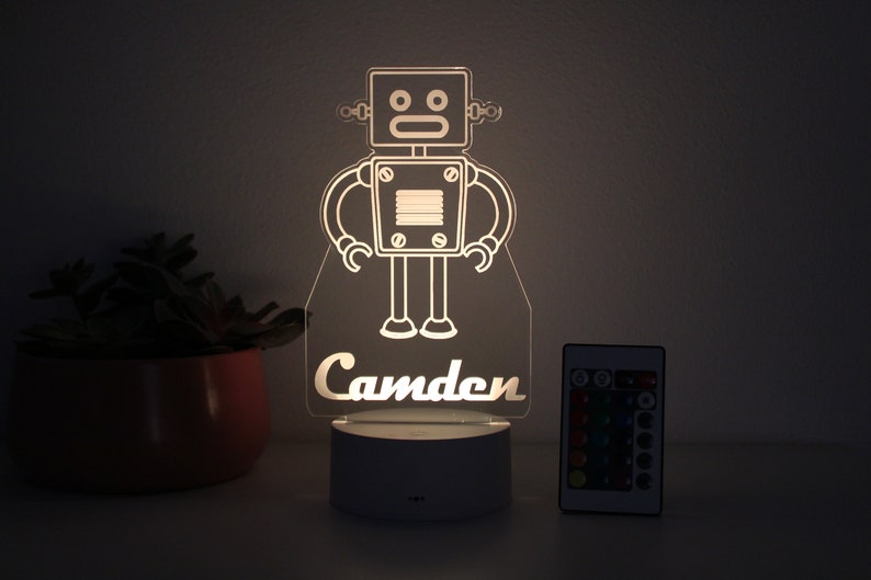May include: A white robot night light with a remote control. The robot has a square head with two eyes and a mouth. The name "Camden" is written below the robot.