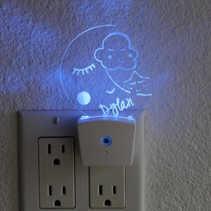 Puede incluir: Una luz nocturna blanca enchufable con un inserto de acrílico transparente que presenta una luna creciente, una nube y una estrella con el nombre "Dylan" escrito debajo. La luz está iluminada en azul.