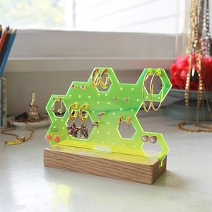 May include: A neon green acrylic earring holder shaped like a honeycomb with a wooden base. The holder has multiple holes for earrings and is filled with various earrings, including gold hoops, star-shaped earrings, and pearl studs.