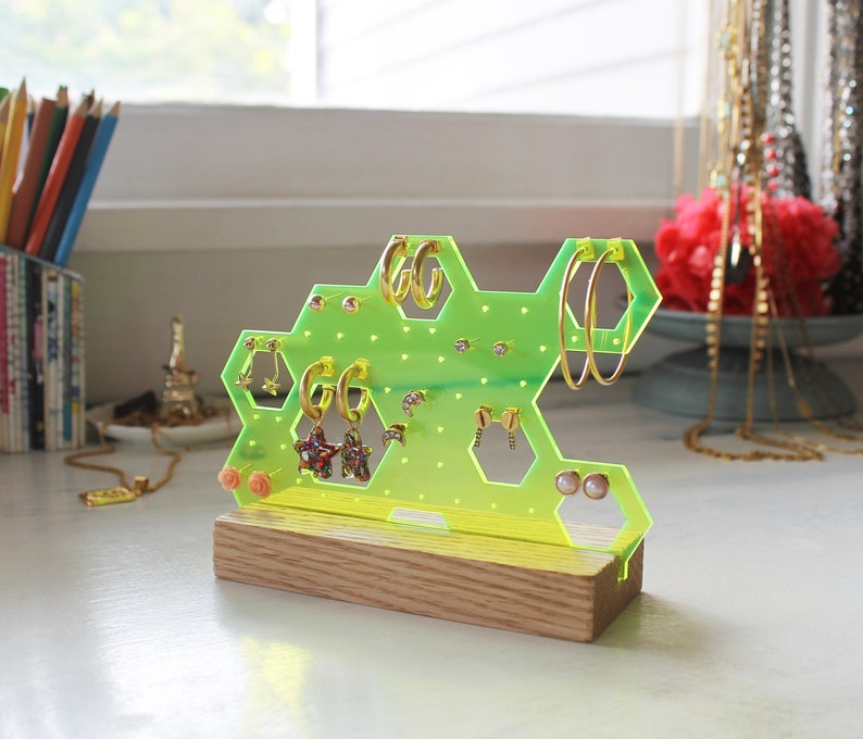 May include: A neon green acrylic earring holder shaped like a honeycomb with a wooden base. The holder has multiple holes for earrings and is filled with various earrings, including gold hoops, star-shaped earrings, and pearl studs.